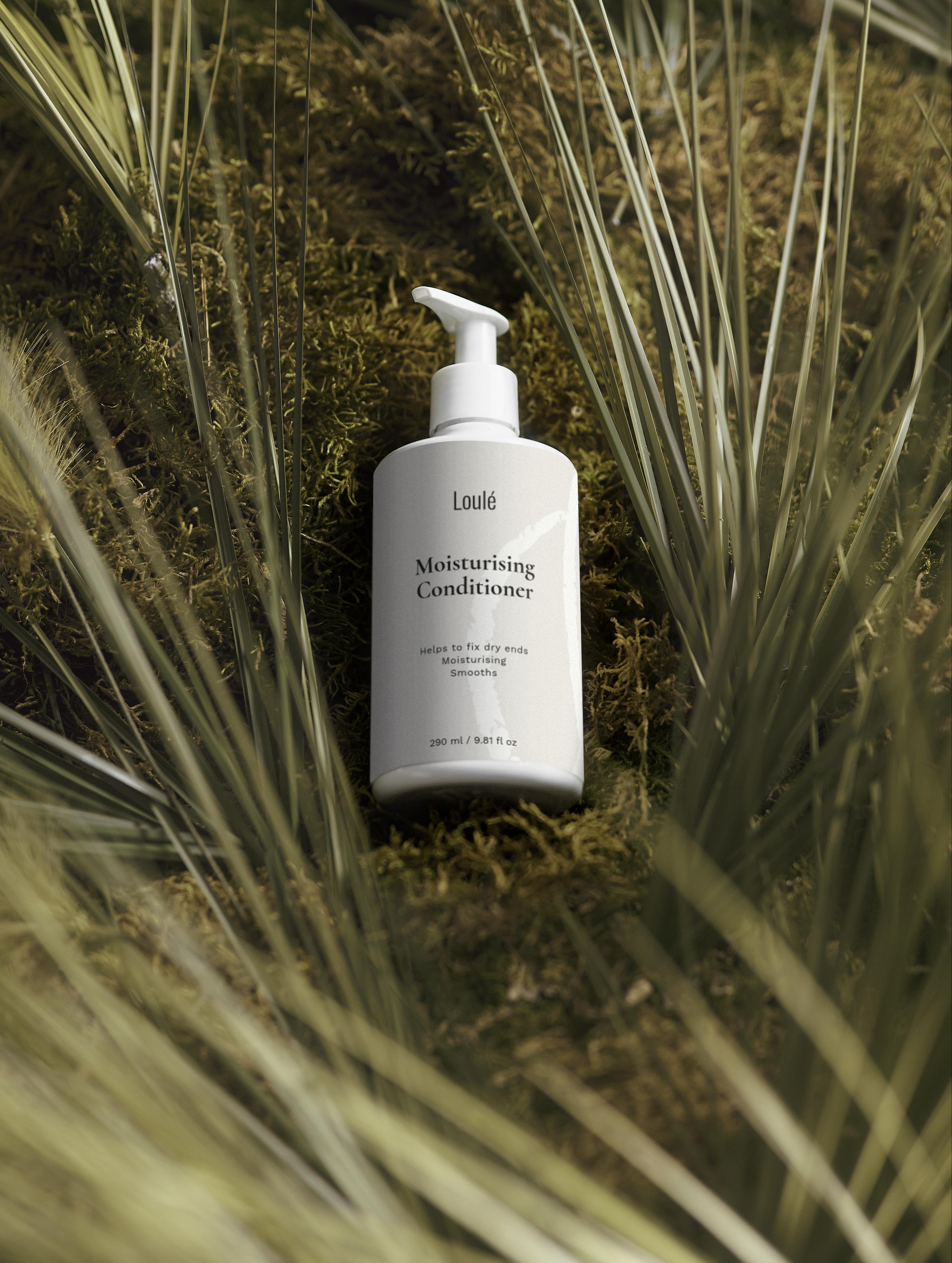 Loulé Moisturising Conditioner bottle with pump, placed on natural moss and surrounded by green grass, ideal for dry ends and lack of volume.