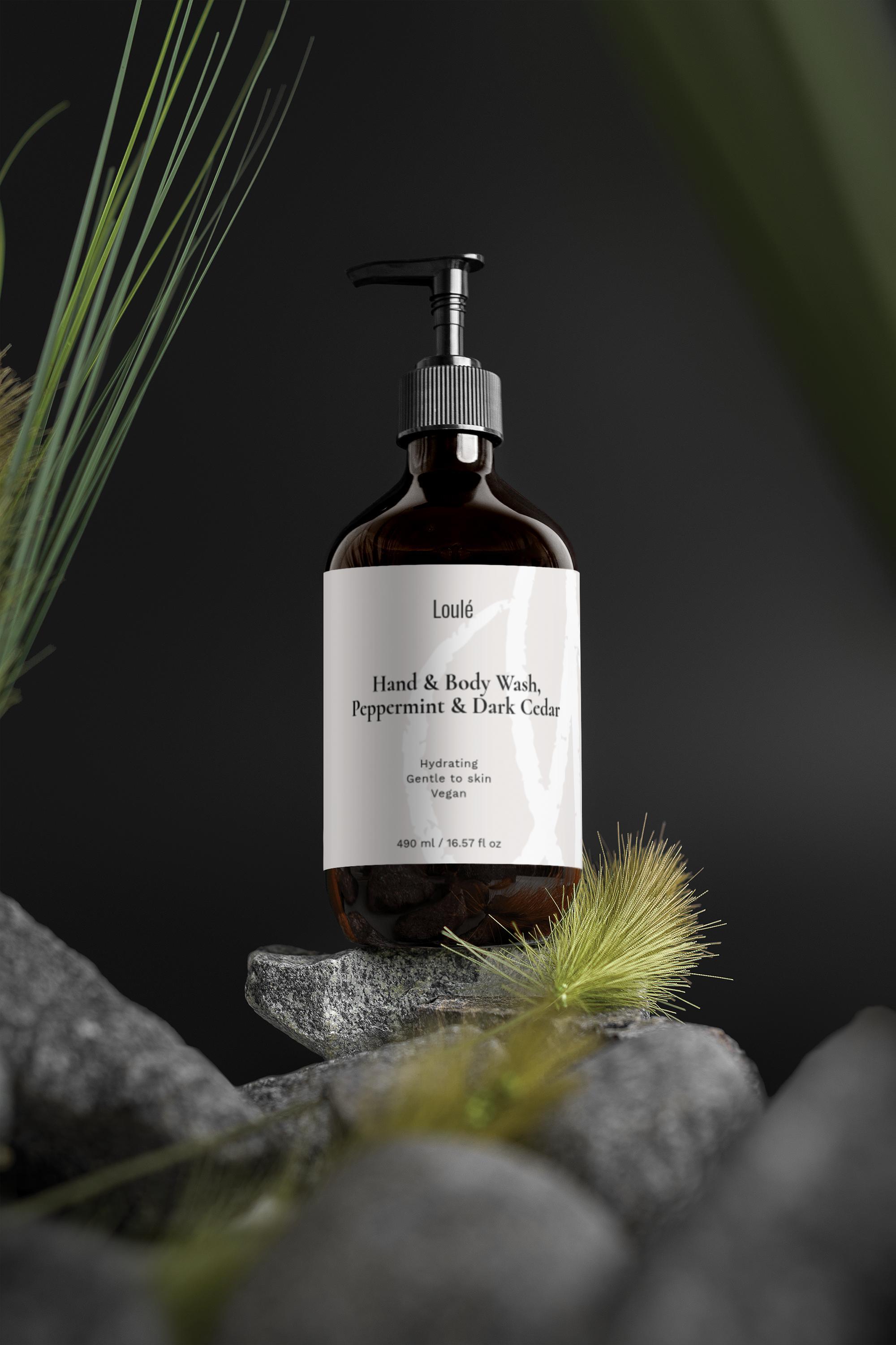 Loulé Skin Hand & Body Wash, Peppermint & Dark Cedar in amber bottle with pump, placed among natural stones and greenery. Hydrating vegan formula for gentle cleansing.